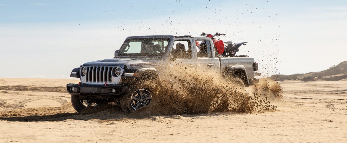 The 2021 Jeep Gladiator Rubicon being driven on sand, carrying a dirt bike in the truck bed.