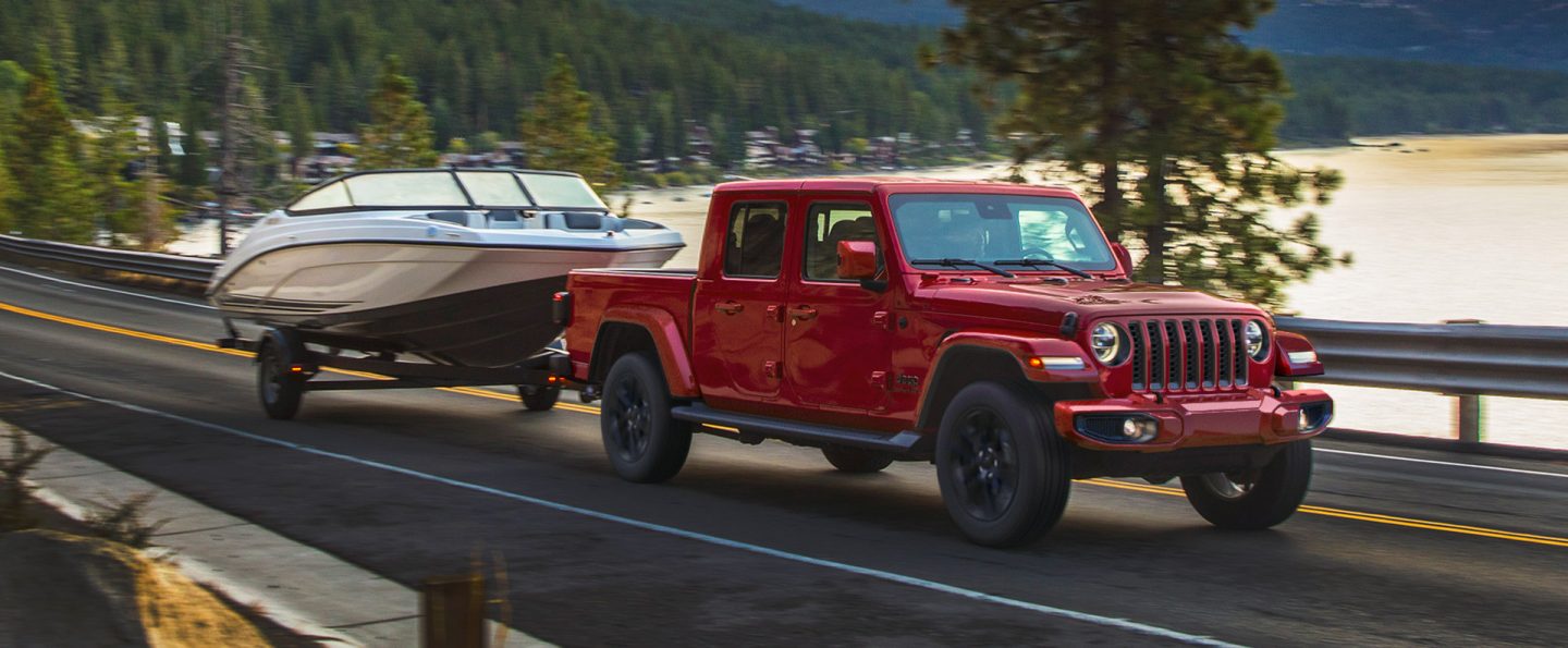 A red 2021 Jeep Gladiator Overland towing a boat beside a lake.